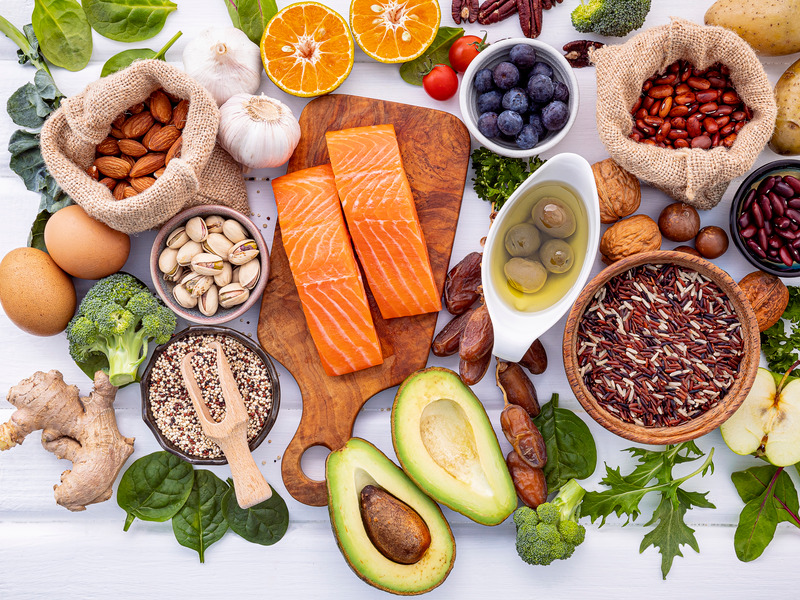 An overhead spread of whole foods &mdash; salmon, avocado, leafy greens, nuts, pulses, berries and wild rice on a pale wooden table.
