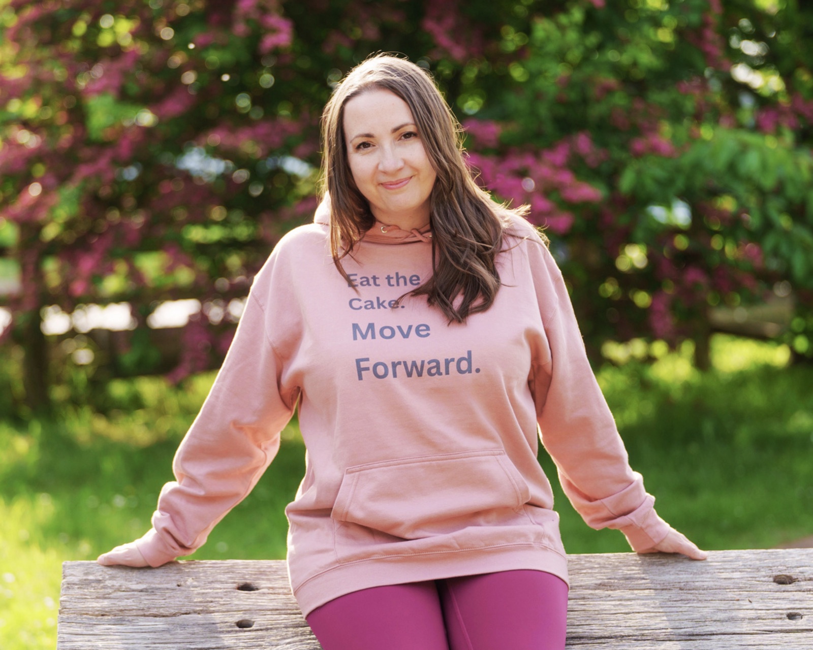 Susan sitting on a wooden bench outdoors, in a pink hoodie reading &ldquo;Eat the Cake. Move Forward.&rdquo;, with blossom behind her.